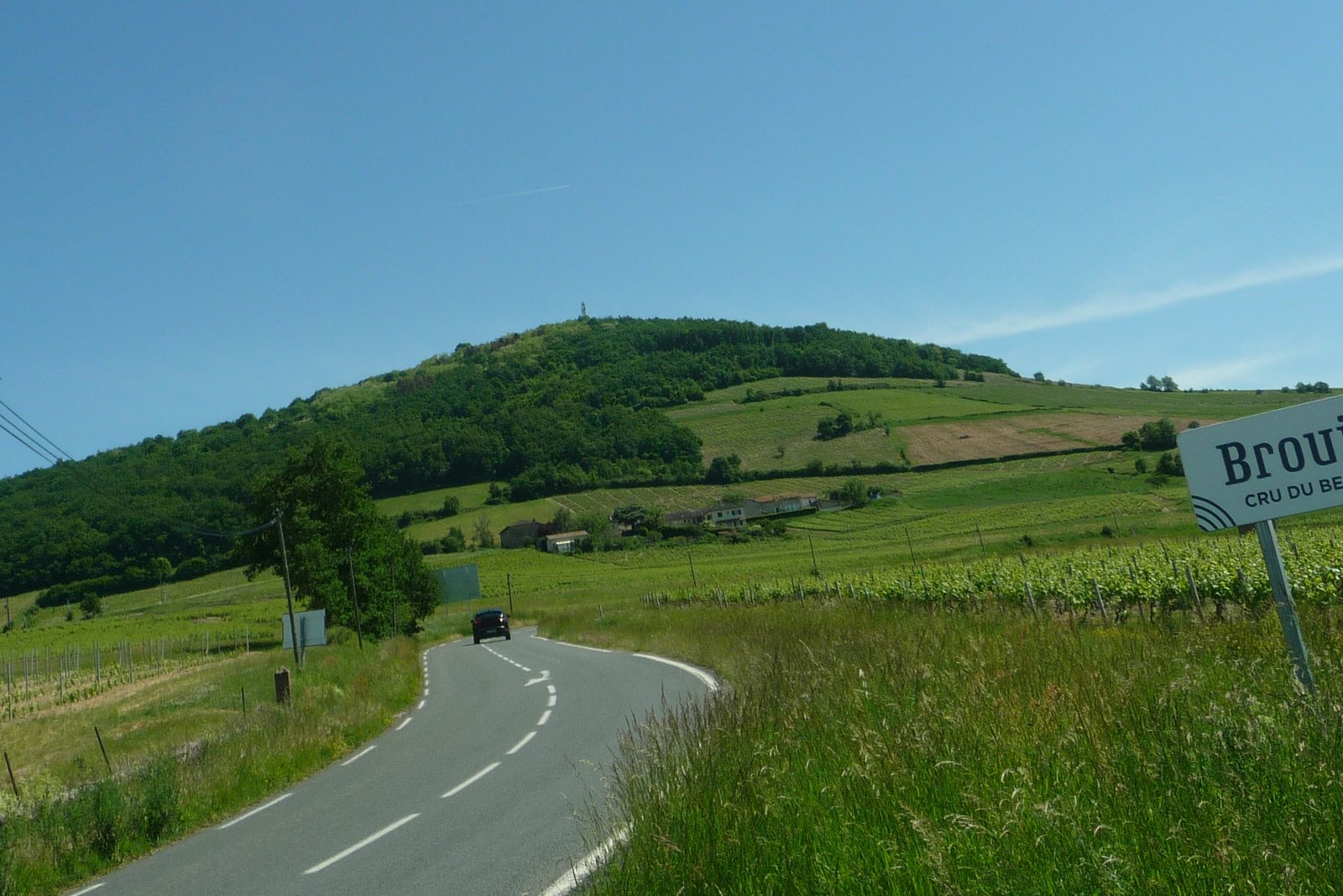 The rounded hill of Mont Brouilly rising beyond vineyards, with a road and a roadside sign reading Brouilly, cru du Beaujolais.